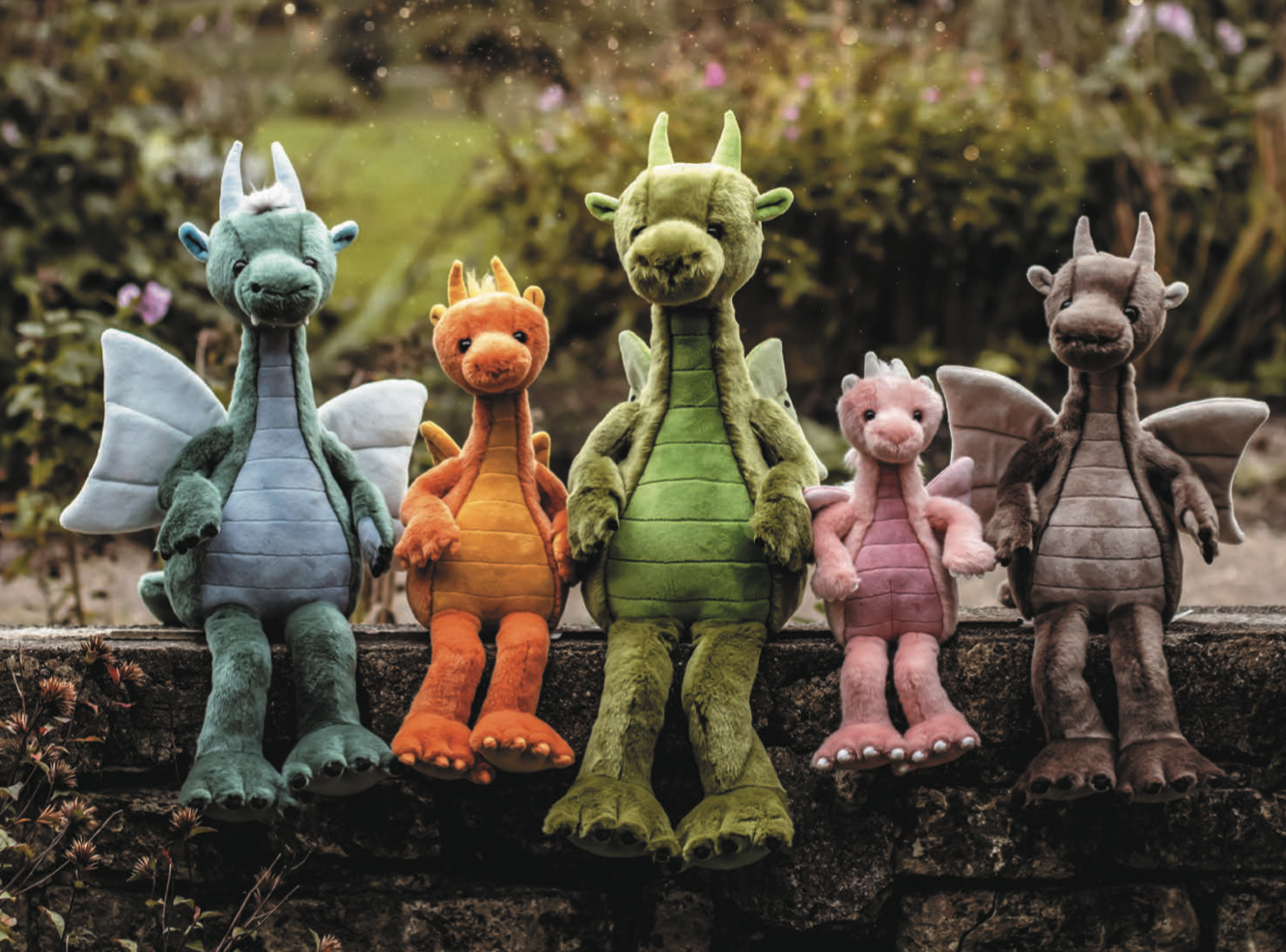 Five colorful dragon plush toys sitting on a stone ledge outdoors, smiling faces, varied sizes, from turquoise to brown.