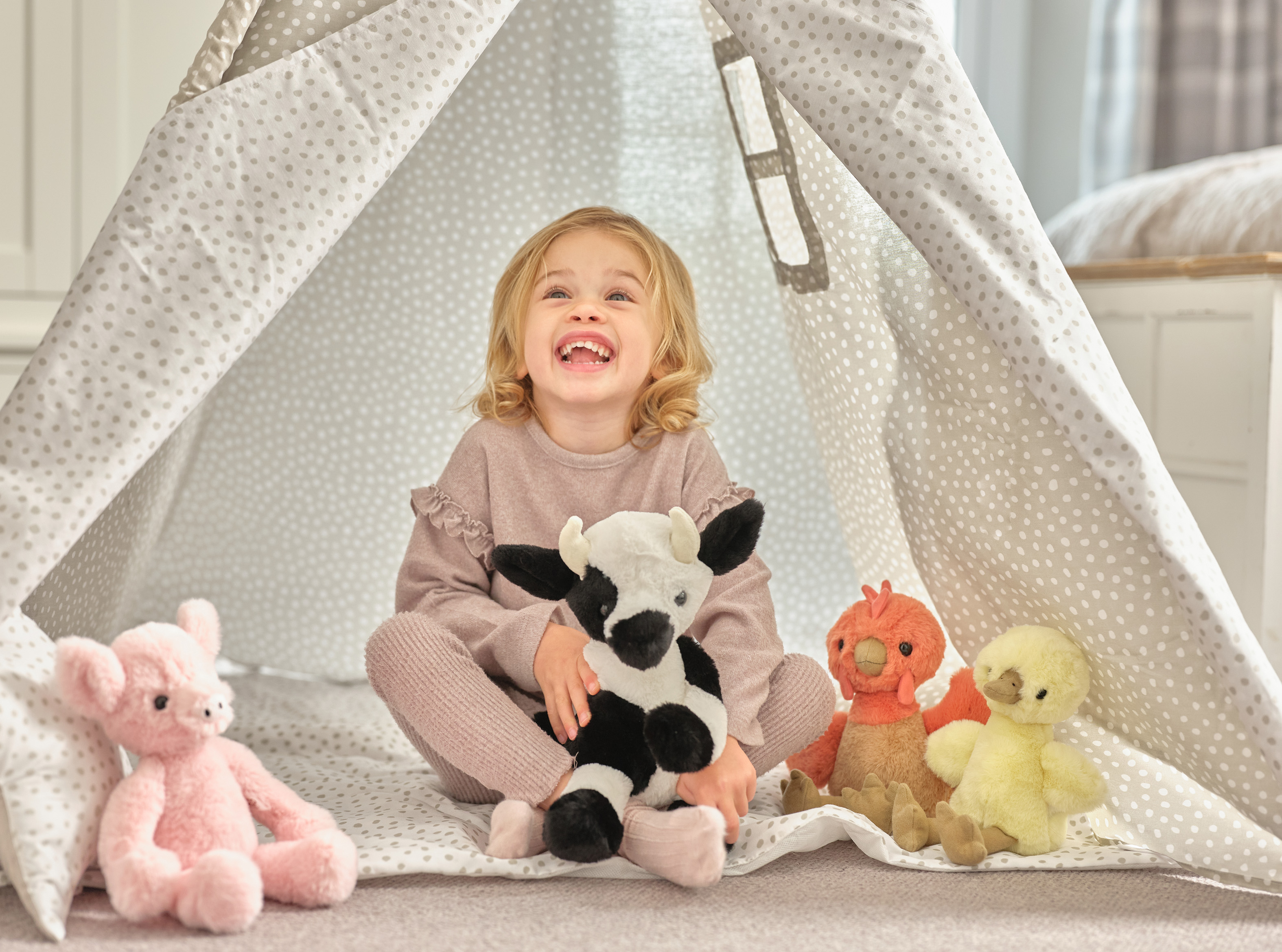A joyful toddler sits inside a polka-dotted fabric play tent, holding a black-and-white plush cow, with pink, orange, and yellow stuffed animals around.