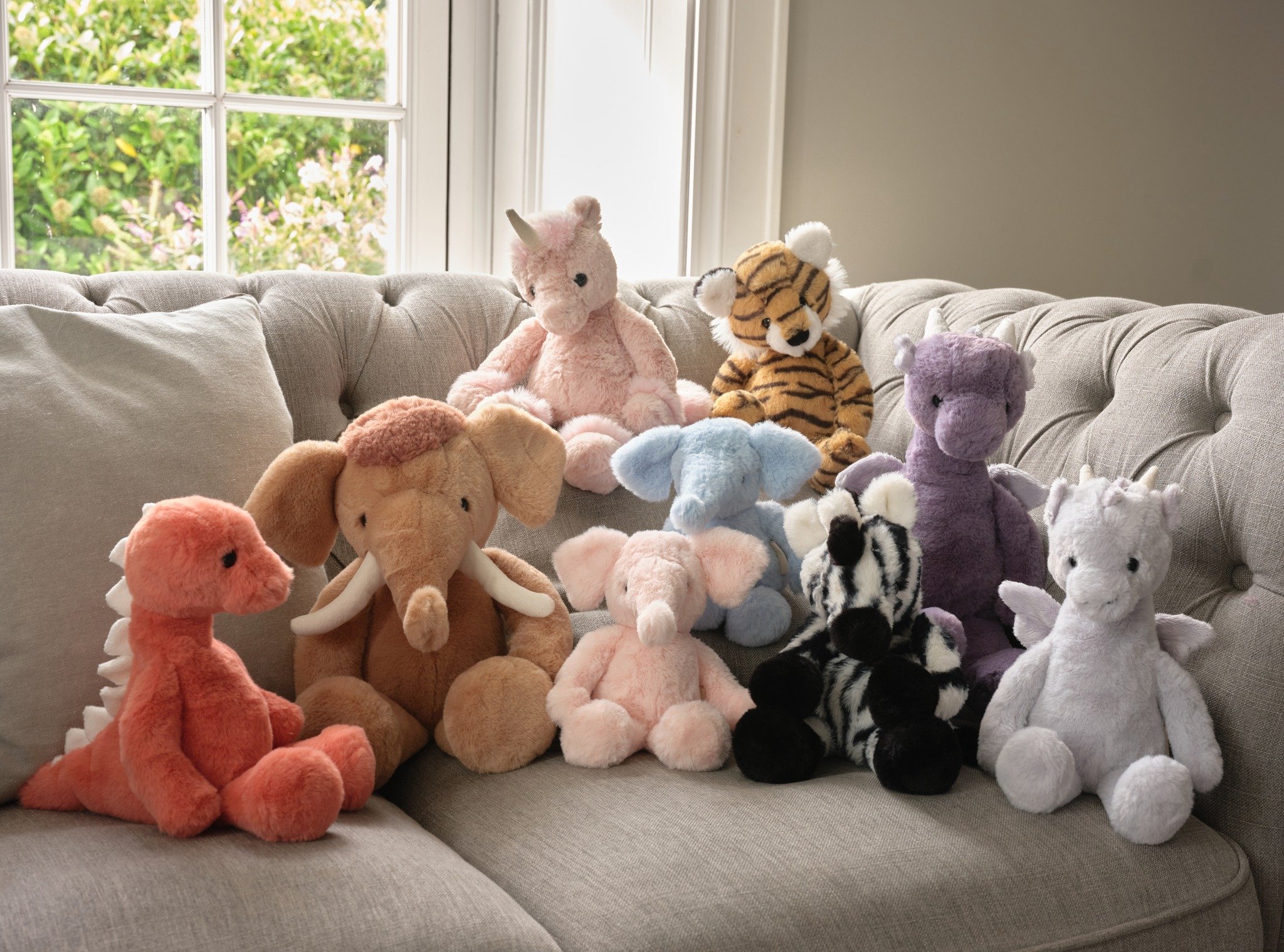 A colourful collection of plush teddy bears and dolls on a light grey sofa by a window.