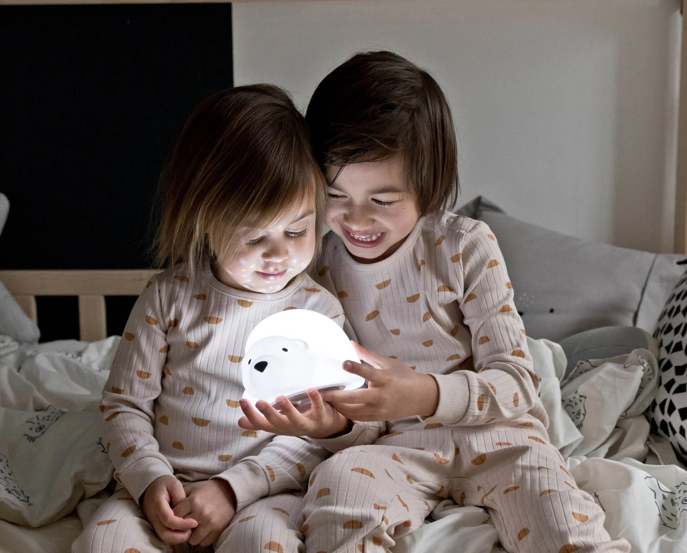 Deux enfants assis sur un lit, souriants, regardent une veilleuse en forme d’ours blanc qui éclaire doucement.