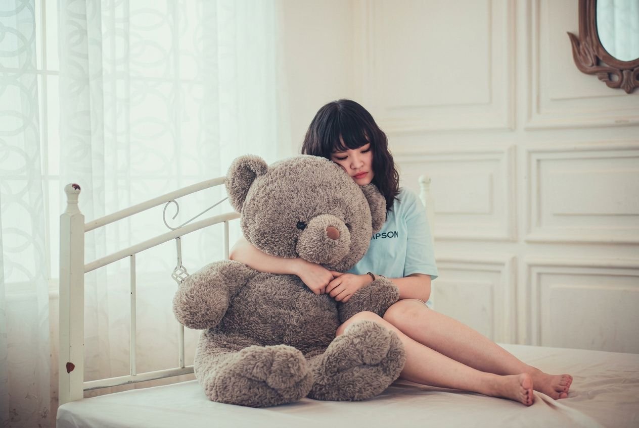 A girl sits on a bed, hugging a large grey teddy bear in a bright, softly lit room.