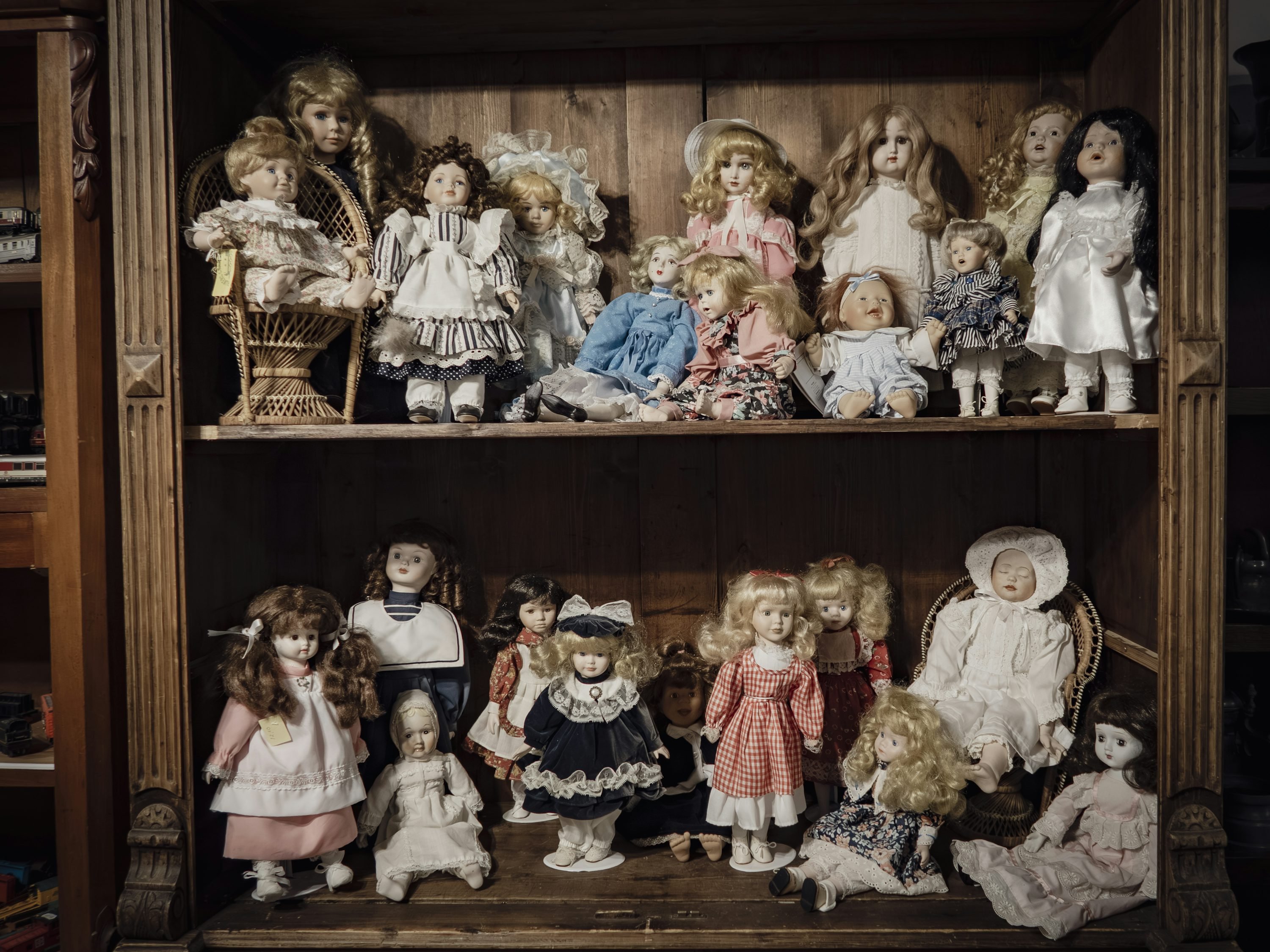 Shelf filled with vintage dolls in period outfits, spanning two levels with wicker chairs and wooden backdrop. Shelf filled with vintage dolls in period outfits, spanning two levels with wicker chairs and wooden backdrop.