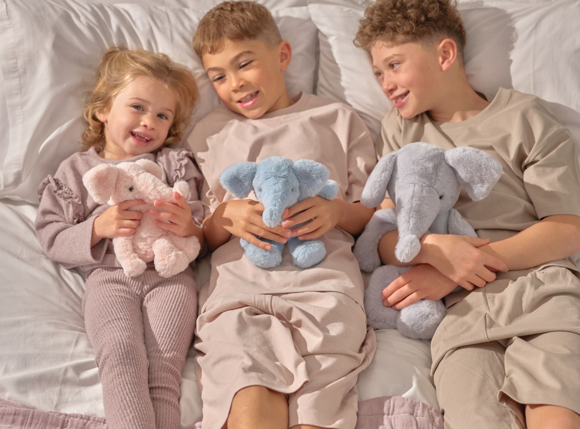 Three children lying in bed, each cuddling a soft teddy bear in pastel colours.