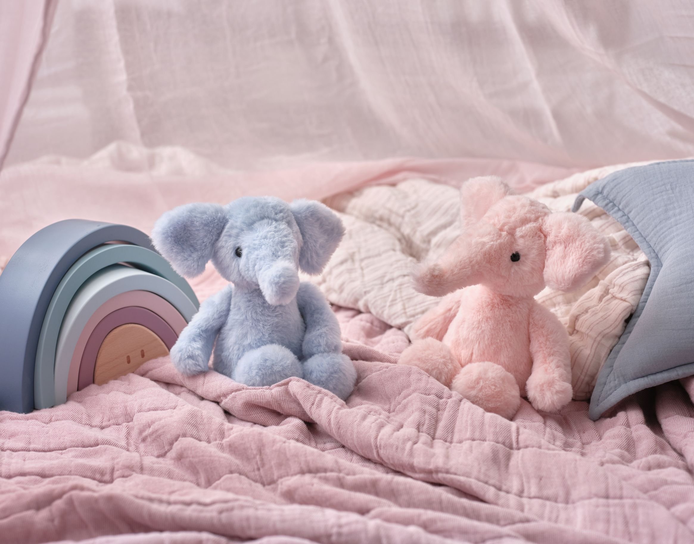Two soft plush elephants, pastel blue and pink, sit on a pink quilted bed with a pale rainbow toy arch and a light blue pillow.