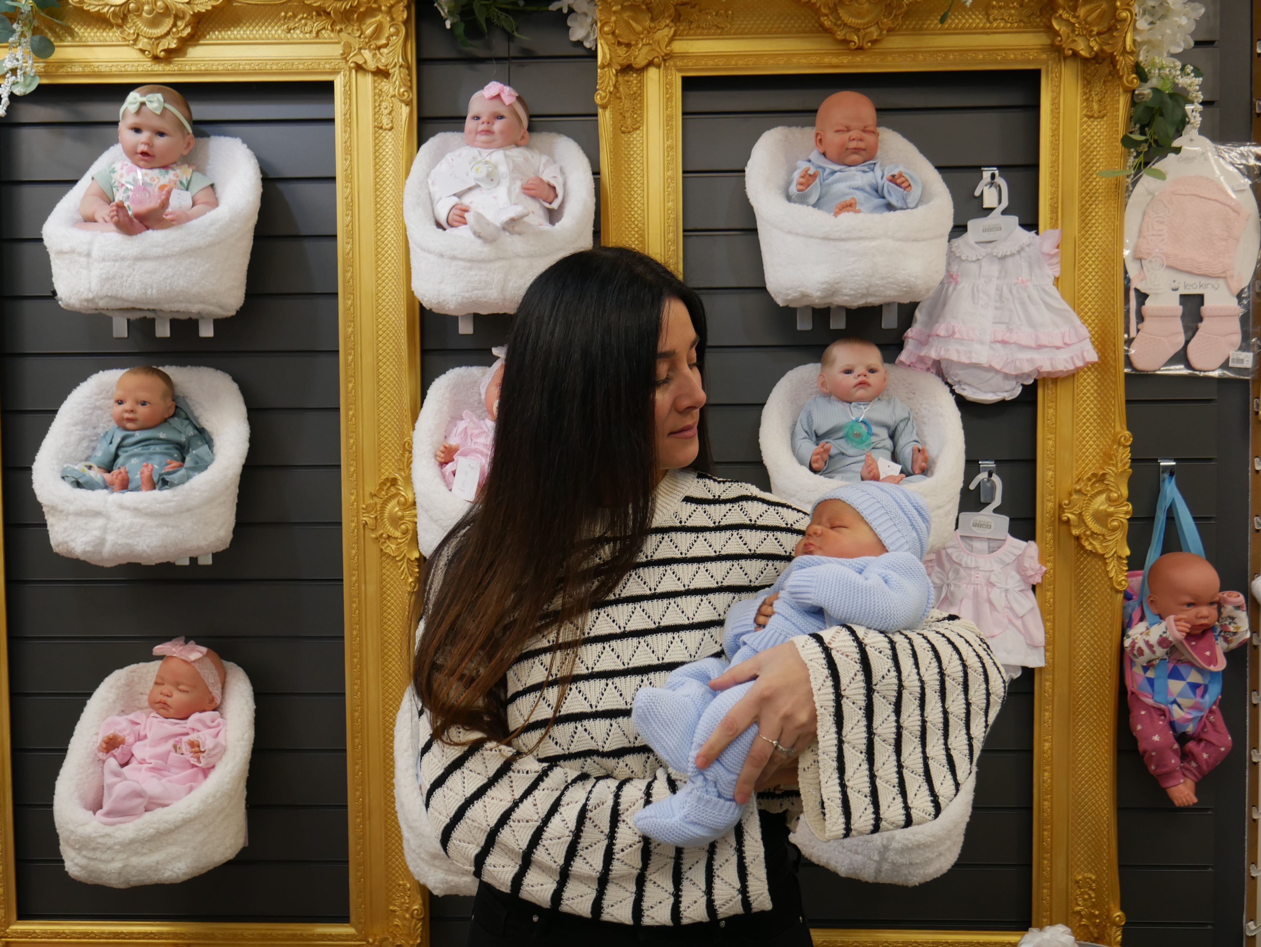 Woman holding a newborn doll in a shop of framed baby doll displays, surrounded by pastel outfits. The setting hints at exploring the psychology of doll collecting.